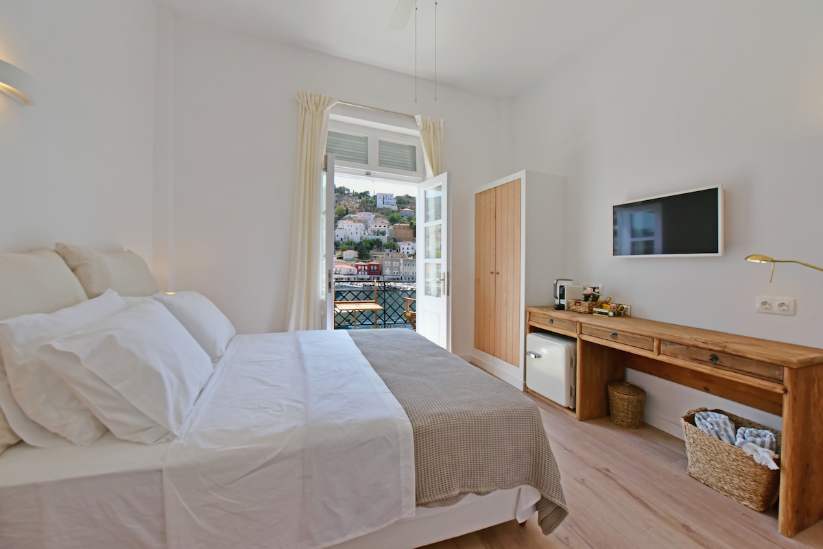 A bright and airy guest room features a spacious bed with a light-colored headboard. Natural light enters through open French doors leading to a shared balcony overlooking the harbor. A wooden console table holds a television and additional amenities, enhancing the room’s functionality.