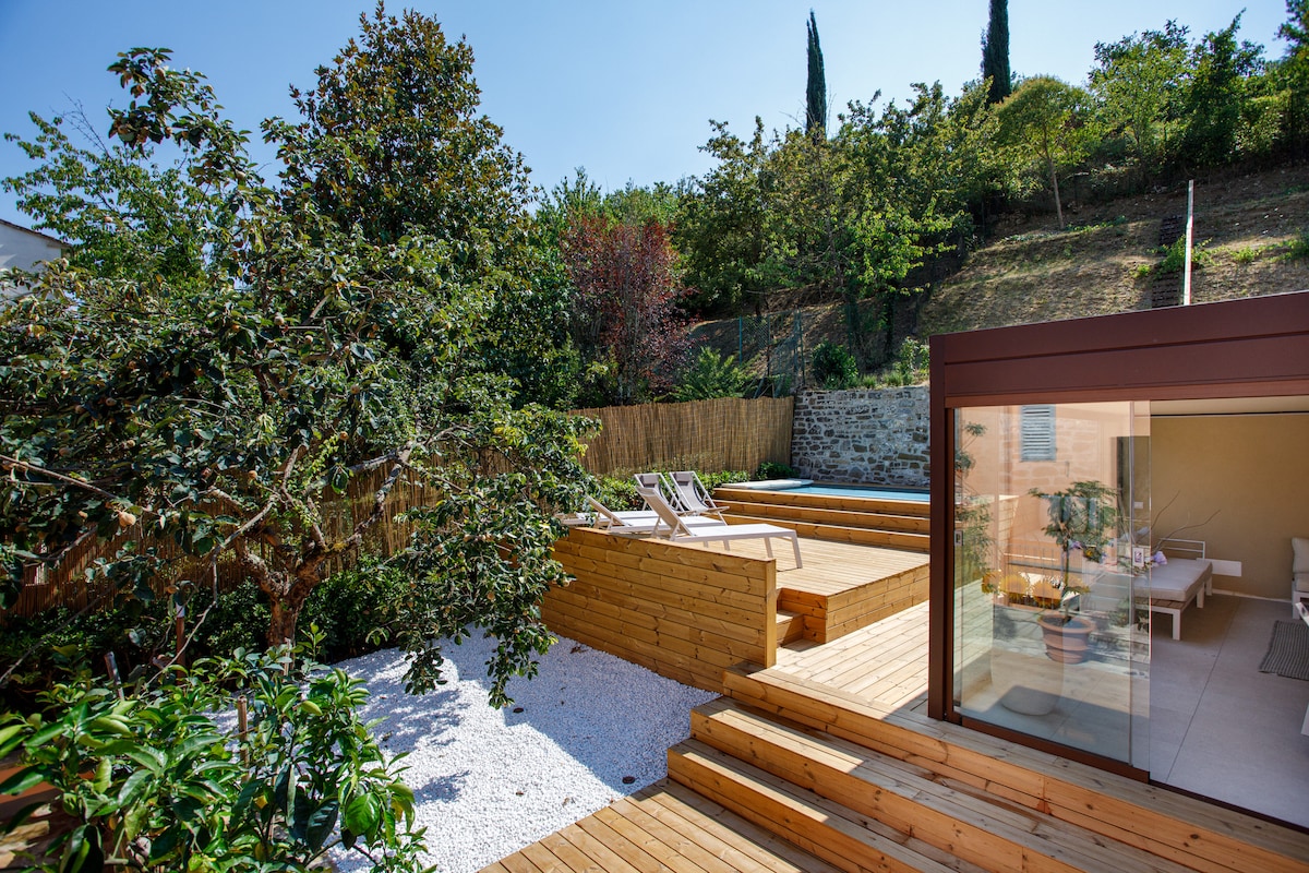 A sunlit outdoor area features wooden decking and lounge chairs, surrounded by trees and greenery. A gravel pathway enhances the natural setting, while the nearby building has large glass doors that provide access to the indoor space.