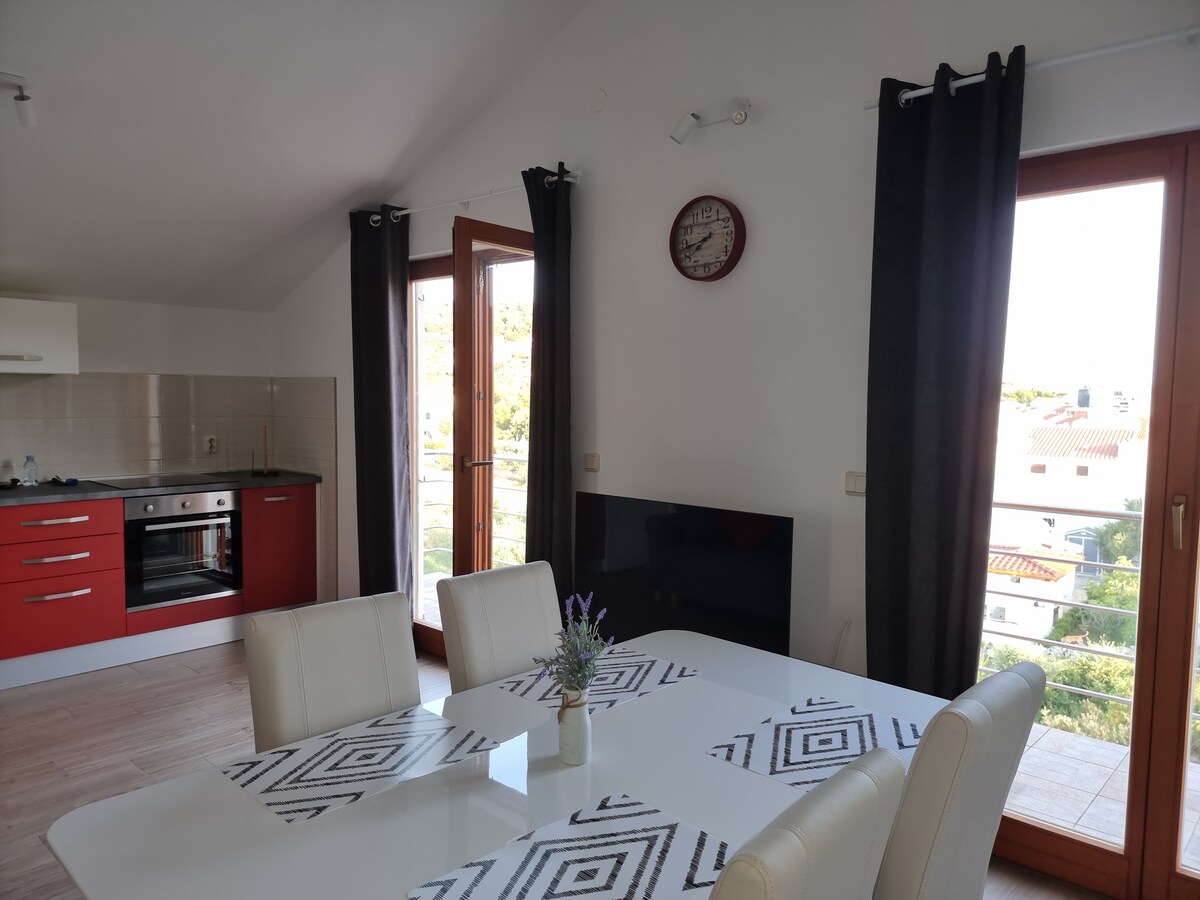 A bright living area is showcased with large windows allowing natural light. A modern dining table accommodates several chairs, enhancing the space. A wall-mounted TV and kitchen area with red cabinetry are visible, along with a stylish clock adorning the wall.