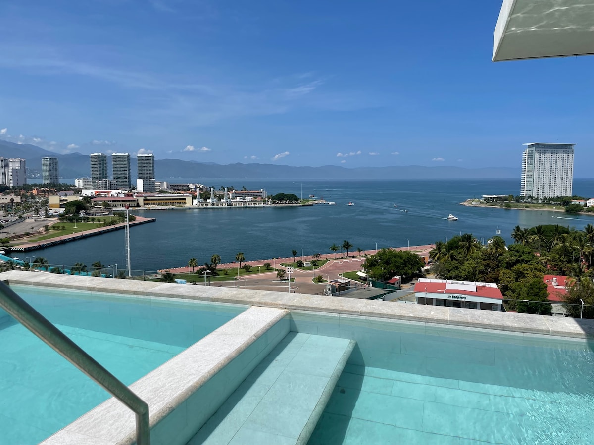 A panoramic view of the coastline is captured from a rooftop infinity pool, with clear blue waters blending into the horizon. The skyline is adorned with modern buildings and lush palm trees, showcasing a vibrant urban landscape against a clear sky.