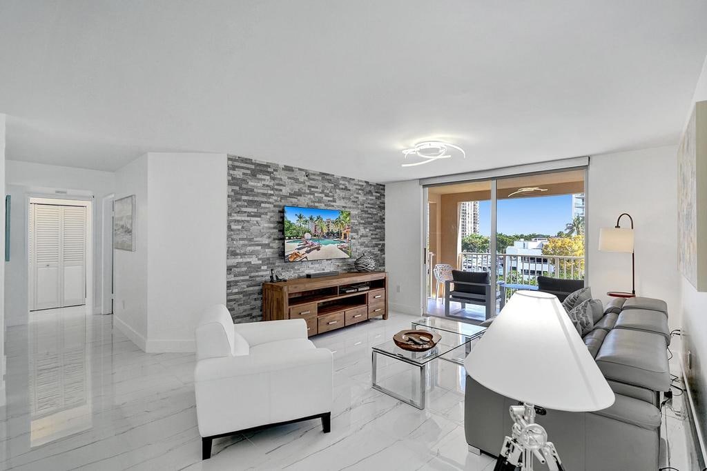 A bright living area features sleek white and gray decor, with a contemporary sofa and armchair positioned around a glass coffee table. A textured feature wall enhances the space, and large sliding doors open to a view of the surrounding marina.