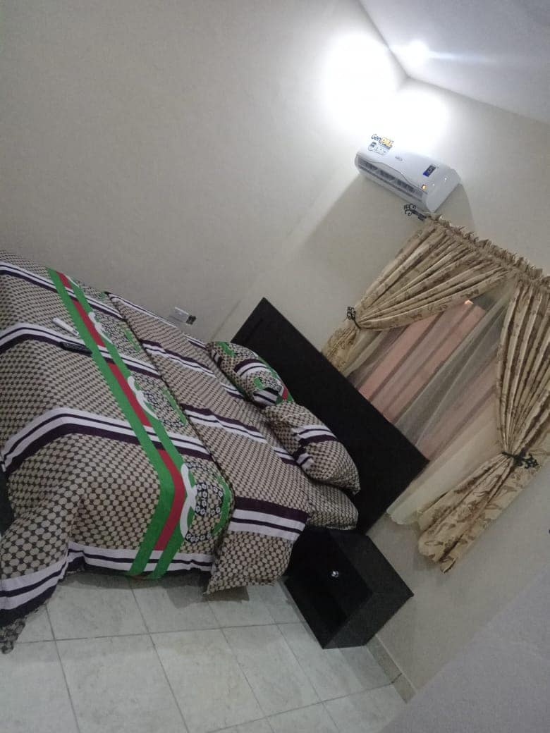A neatly arranged sleeping area is featured, with a spacious bed covered in patterned bedding. A bedside table stands beside the bed, while a window adorned with long curtains allows natural light to enter. An air conditioning unit is mounted above for comfort.