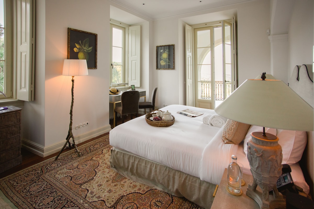 The elegantly furnished suite features a spacious bed with crisp white linens, complemented by an area rug. Natural light floods the room through large windows, alongside tasteful artwork on the walls. A lamp and a decorative table add to the room's charm.