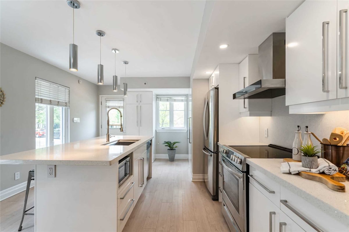The kitchen features modern cabinetry and stainless steel appliances, including a refrigerator and stove. A large island with bar seating provides additional workspace. Natural light floods the area through a nearby window, while a potted plant adds a touch of greenery.