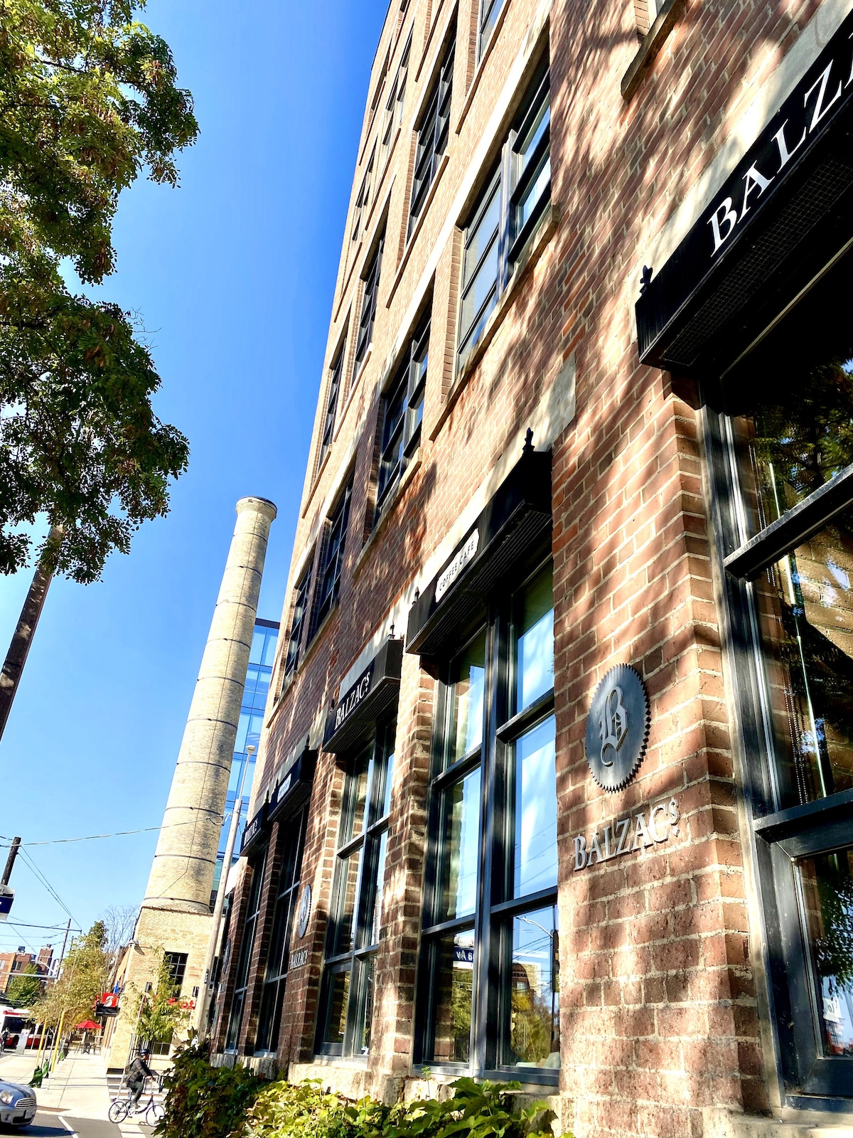 A brick building is shown with large windows reflecting sunlight. The structure features a historical chimney nearby, hinting at the area's industrial past. The scene is framed by tree branches, adding greenery to the urban setting.