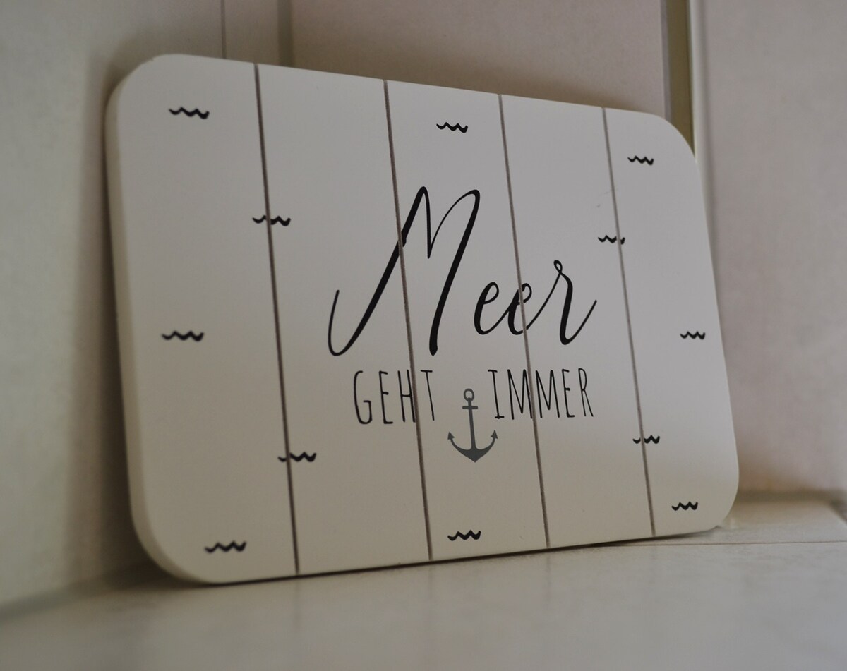 A decorative wooden sign is displayed against a light background. The sign features the word 'Meer' scripted in an elegant font, overlaying a design of horizontal lines and nautical motifs, imparting a sense of coastal charm.