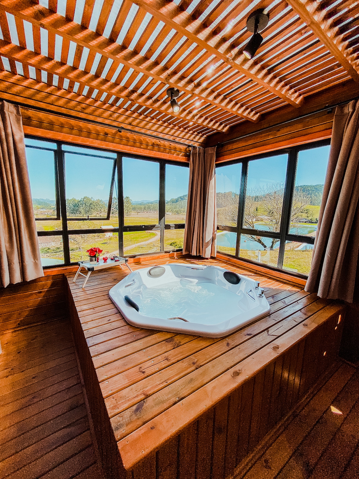 A welcoming hot tub is positioned on a wooden deck, surrounded by large windows that provide ample natural light. The view includes lush greenery and distant hills. Soft curtains frame the windows, creating a serene atmosphere.