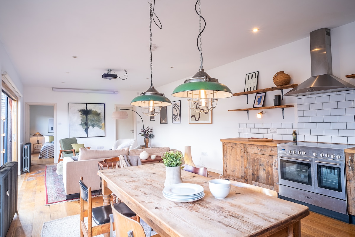 An open-plan living space features a large wooden dining table surrounded by dark chairs. Pendant lights hang above, illuminating the area. A modern kitchen with stainless steel appliances and open shelving is present. A cozy seating area is visible in the background, with light-colored furniture and art on the walls.