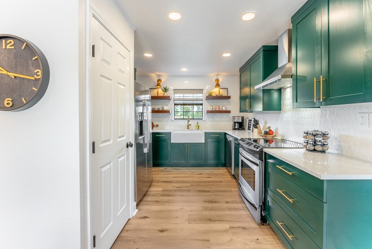 A fully equipped kitchen is presented with deep green cabinetry and modern stainless steel appliances. A large window allows natural light to illuminate the work areas. Decorative elements, including plants and placed items, add a touch of charm. A clock is visible on the wall.