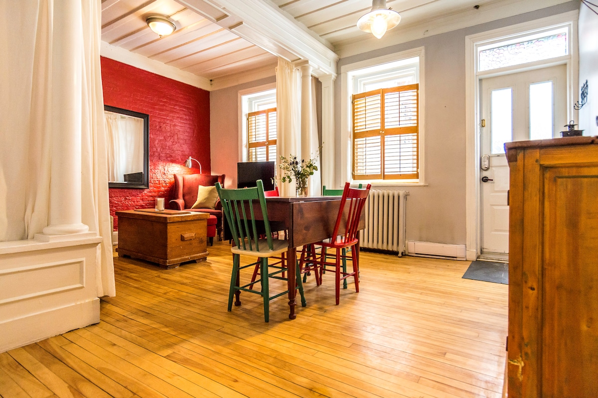 An open living area features a wooden floor and a dining table surrounded by colorful chairs. A red accent wall contrasts with light grey walls. A comfortable seating area is visible, along with a window dressed in wooden shutters that allows natural light to enter.