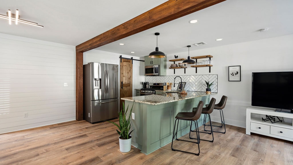A modern kitchen is showcased with a sizable island featuring three contemporary bar stools. Stainless steel appliances, including a refrigerator and oven, are visible. Smooth wood floors complement the light walls, and decorative shelving adds charm above the countertop. A small plant adds a touch of greenery.