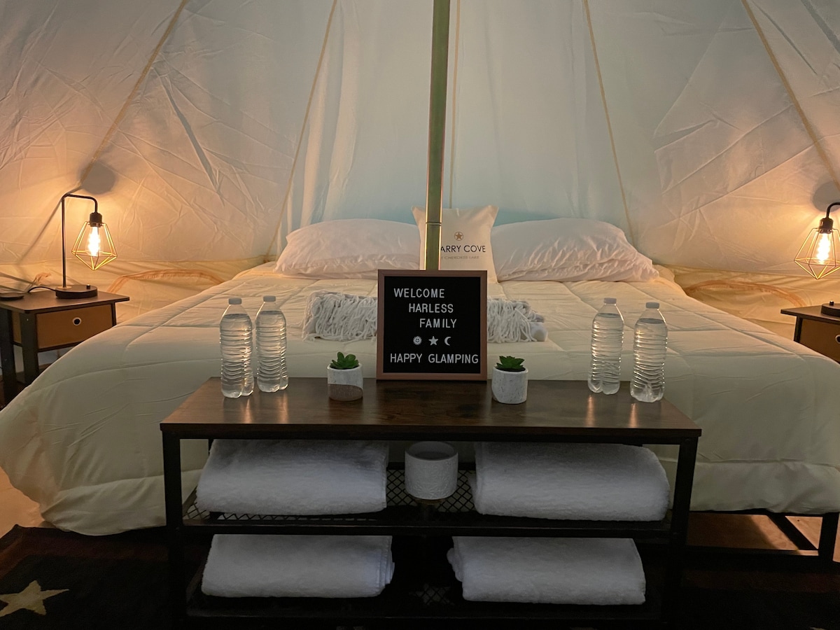 A luxury bell tent interior displays a spacious bed with crisp linens and a decorative pillow. A wooden bench holds neatly stacked towels and water bottles. Soft lighting is provided by lamps on both sides of the bed. A welcoming sign enhances the relaxed ambiance.