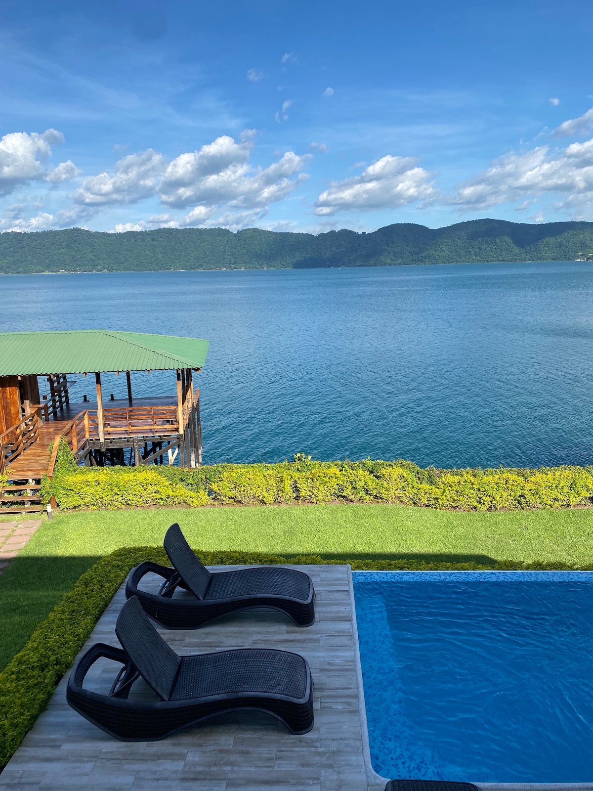 A serene view of a calm body of water is captured, with lush mountains in the background. Two lounge chairs are positioned beside a swimming pool, creating a relaxing outdoor space. An overwater dock is visible to the left, adding to the tranquil atmosphere.