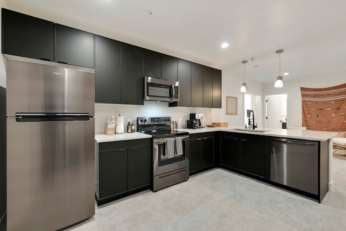 A sleek kitchen features stainless steel appliances, including a refrigerator and a microwave. Dark cabinetry contrasts with light countertops. Under-cabinet lighting enhances the workspace, while a dining area is visible in the background, showcasing a modern aesthetic.