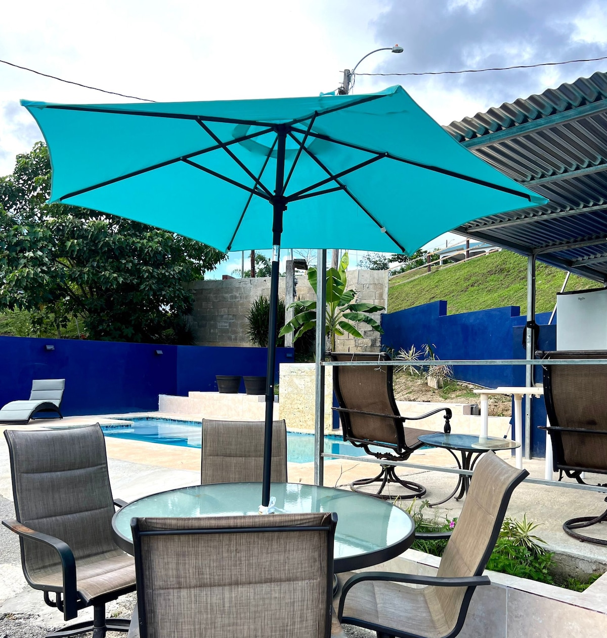 A round glass-top table is surrounded by four light wicker chairs, each featuring a cushioned seat. Above, a large turquoise umbrella provides shade. In the background, a private pool is visible, complemented by a vibrant blue wall and greenery.