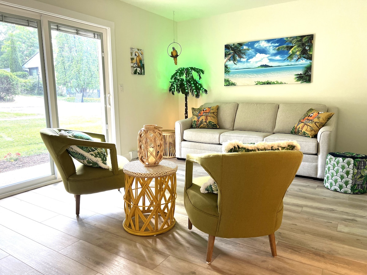 A bright living area features a light-colored sofa and two green accent chairs. A round, woven coffee table sits at the center, complemented by decorative pillows on the sofa. Large sliding doors open to a view of the outdoors, enhancing the space's natural light.