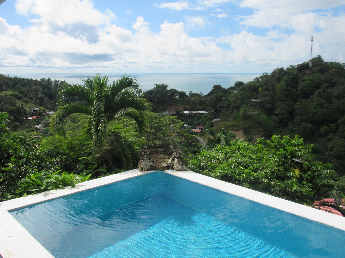 An inviting pool is positioned at the edge of the property, featuring clear blue water. Lush greenery surrounds the area, and ocean views are visible in the distance under a partly cloudy sky. The tranquil setting offers a sense of privacy and relaxation.