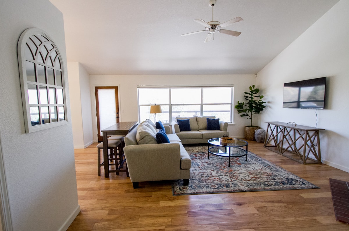 A bright living area features a comfortable sofa and a round coffee table centered on a decorative rug. Large windows allow natural light to fill the space, complemented by a mounted flat-screen TV and a tall plant in the corner, enhancing the room's inviting design.