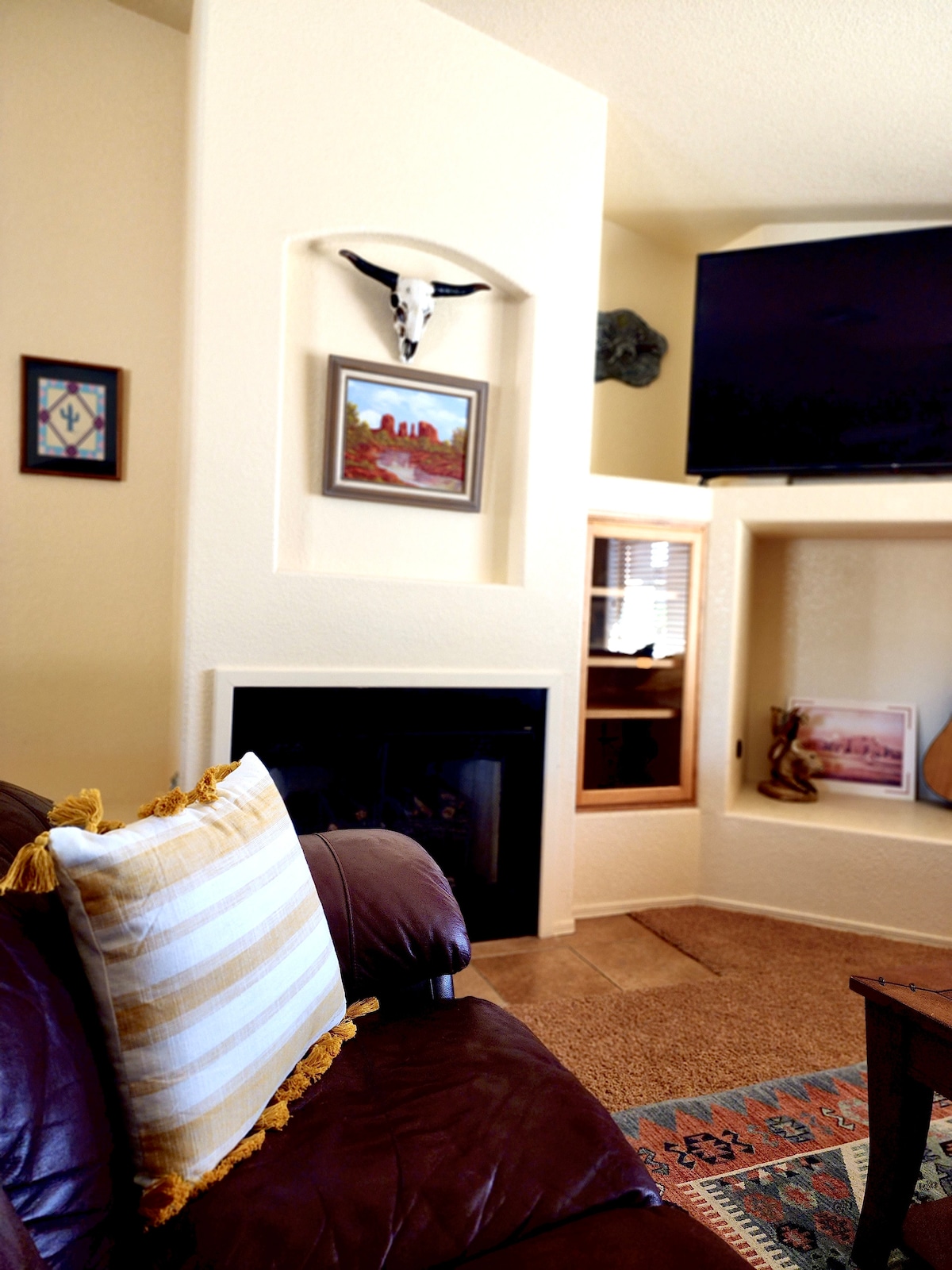 A cozy living space is highlighted by a leather couch adorned with a striped pillow. A wall-mounted television sits above a decorative fireplace, while framed artwork and a small display unit add character to the warm, inviting room.