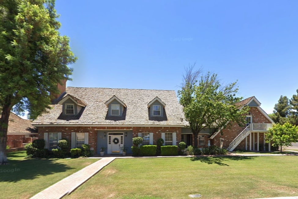 The exterior of a charming red brick home is shown, featuring a steeply pitched roof and large front yard. A separate upstairs apartment with a private staircase can be seen on the right, surrounded by mature trees and neatly manicured shrubbery.