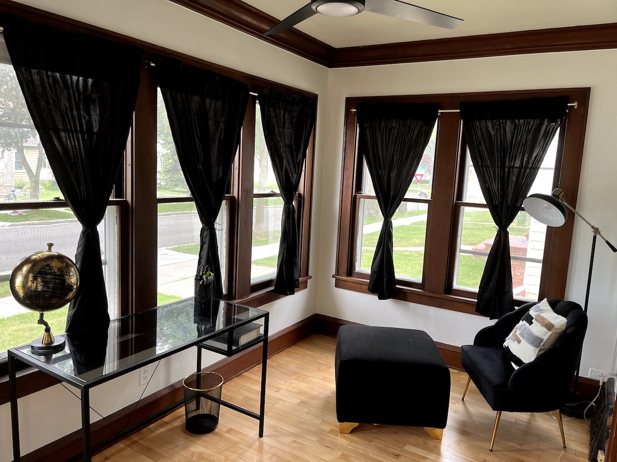 A sunroom is featured with large windows framed by dark curtains, allowing for natural light. A sleek glass table is positioned next to a modern black chair with an accent pillow. A minimalist floor lamp and a decorative globe enhance the space.