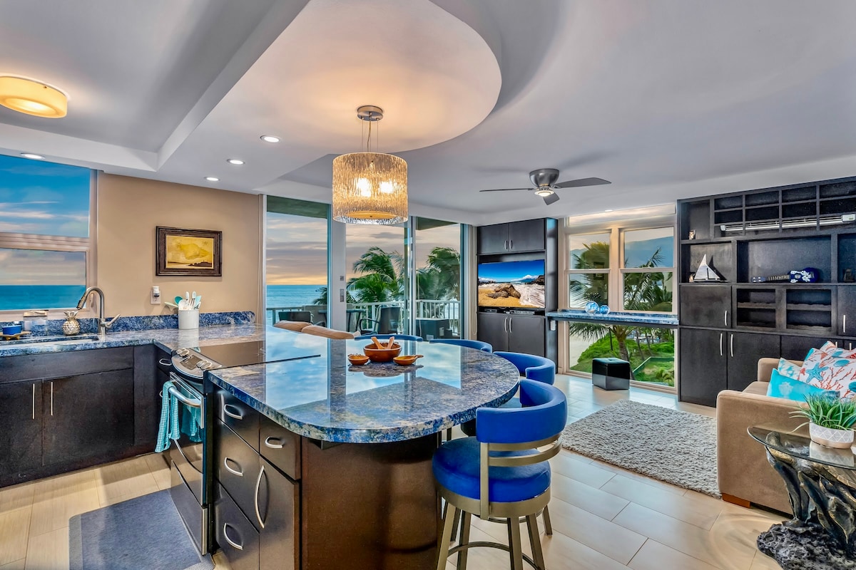 A modern kitchen is characterized by dark cabinetry and a large island with a blue granite countertop. Soft lighting is provided by a chandelier above. Large windows allow views of the ocean and sunset, enhancing the light and airy feel of the space.