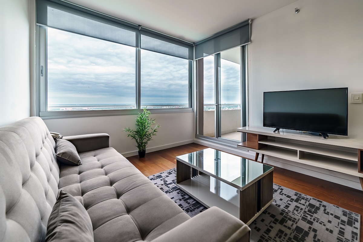 A modern living area features a spacious gray sofa and a glass coffee table situated on a patterned rug. A flat-screen TV is positioned on an elegant console. Large windows provide a view of the outside, framed by light curtains.