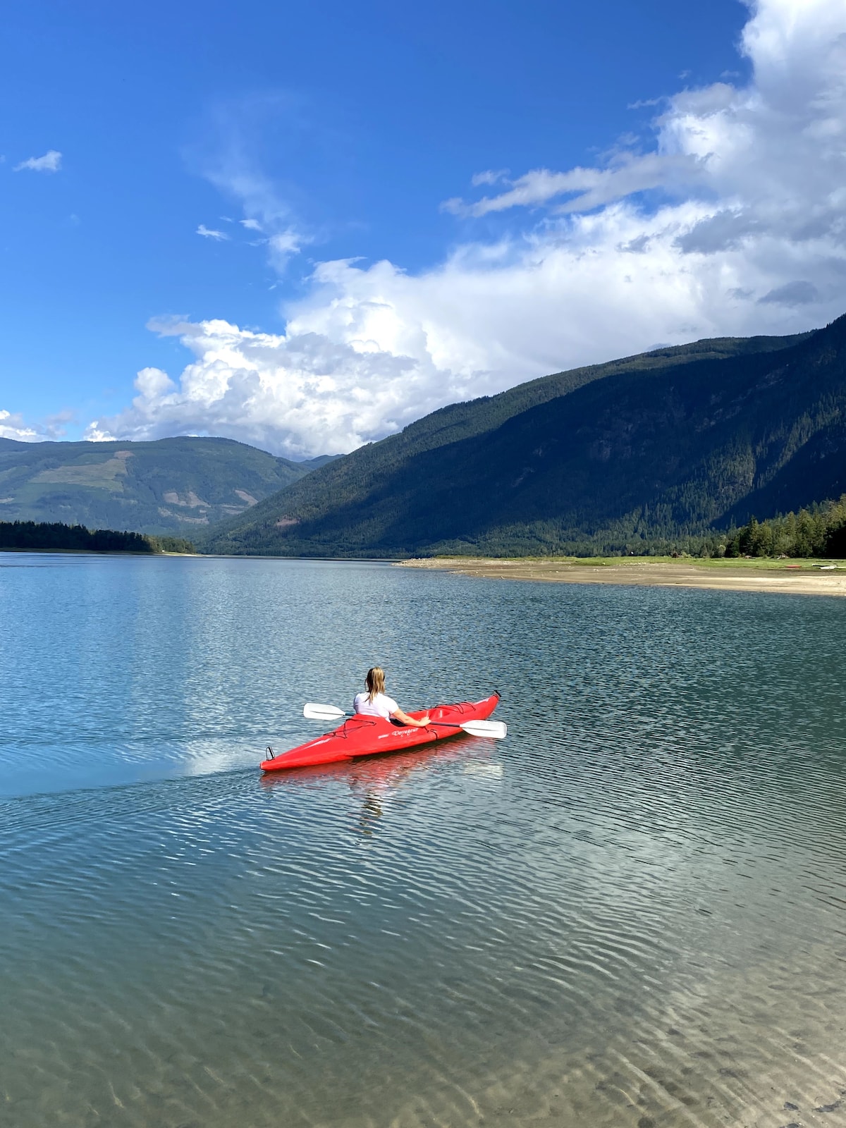 A person is seen kayaking in a bright red kayak on calm waters, surrounded by lush green mountains. The sky is partly cloudy, and sunlight reflects off the water, creating a tranquil setting. The shoreline is visible in the background, indicating a spacious environment.