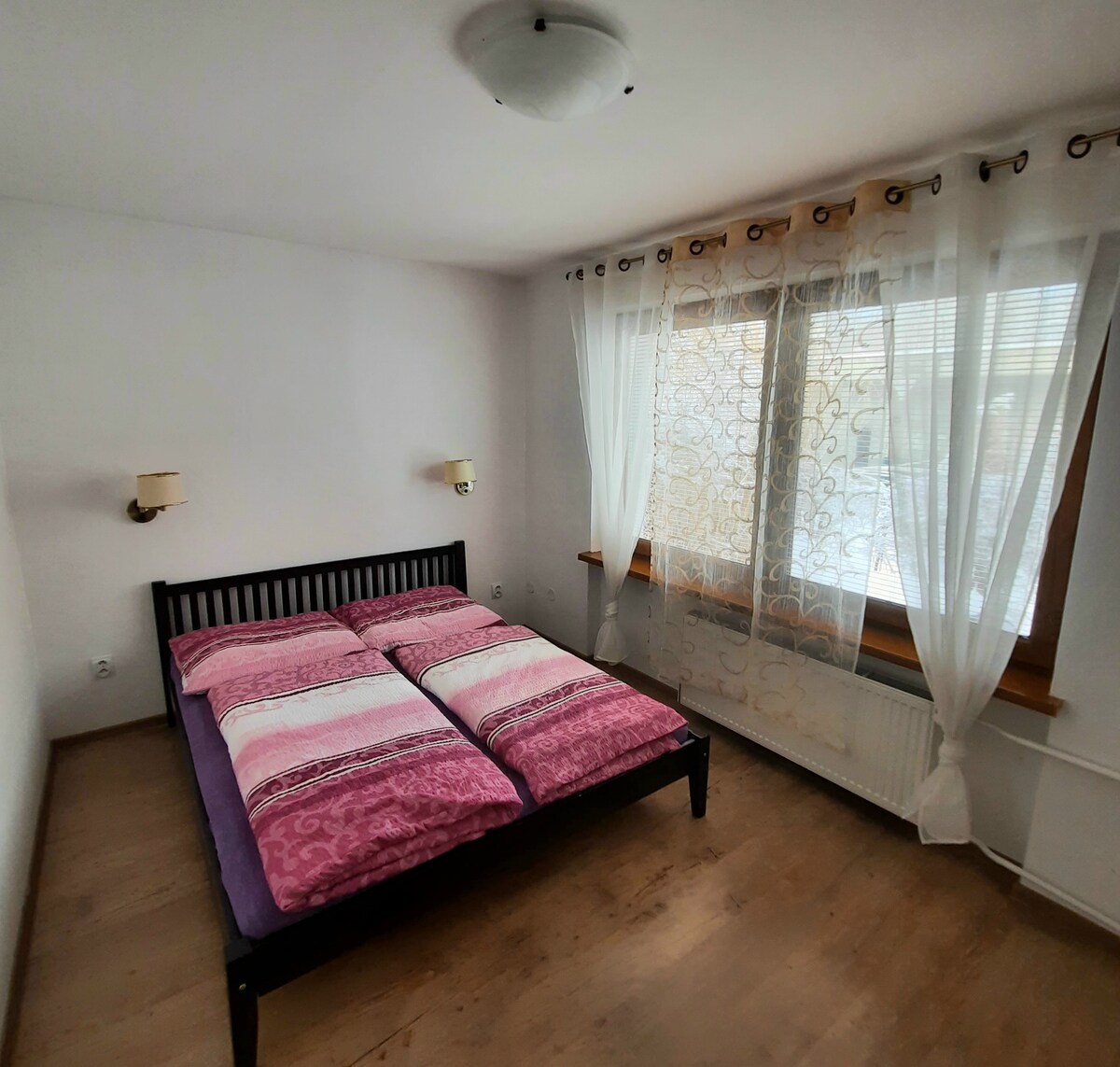 A comfortable bedroom features a spacious bed with pink and white bedding. Natural light filters through sheer curtains, illuminating the room. Wall sconces provide additional lighting, complementing the light-colored walls and wooden flooring.