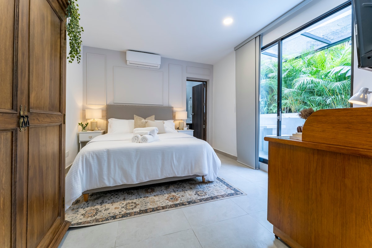 A spacious bedroom is highlighted by a king-size bed topped with crisp white linens. Natural light enters through glass sliding doors, revealing greenery outside. An antique wooden wardrobe and a wooden desk accent the room, contributing to a calm and inviting atmosphere.