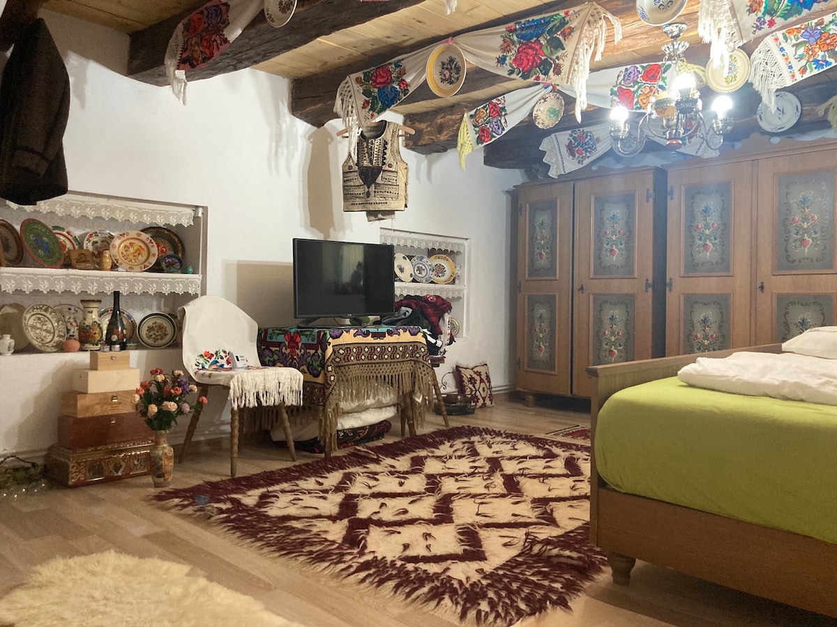 A cozy living area features a colorful area rug on polished wood flooring. A vintage-style wooden cabinet with intricate designs stands beside a television. Decorative plates adorn the shelves, while traditional textiles drape from the ceiling, creating a warm, culturally rich ambiance.