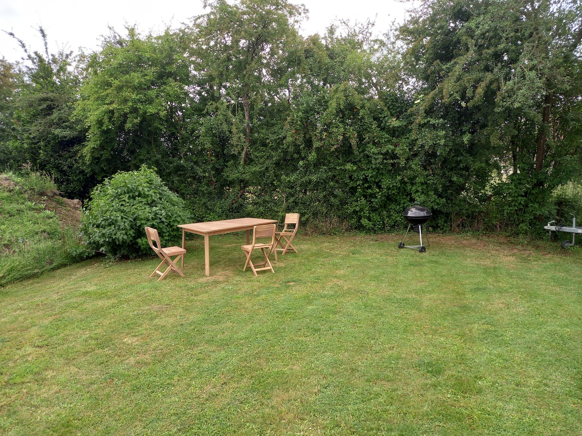 A sunny garden space is presented, featuring a wooden table surrounded by three folding chairs. A charcoal barbecue grill is positioned nearby, with lush green shrubs creating a natural backdrop. The well-maintained lawn area offers ample space for outdoor activities.