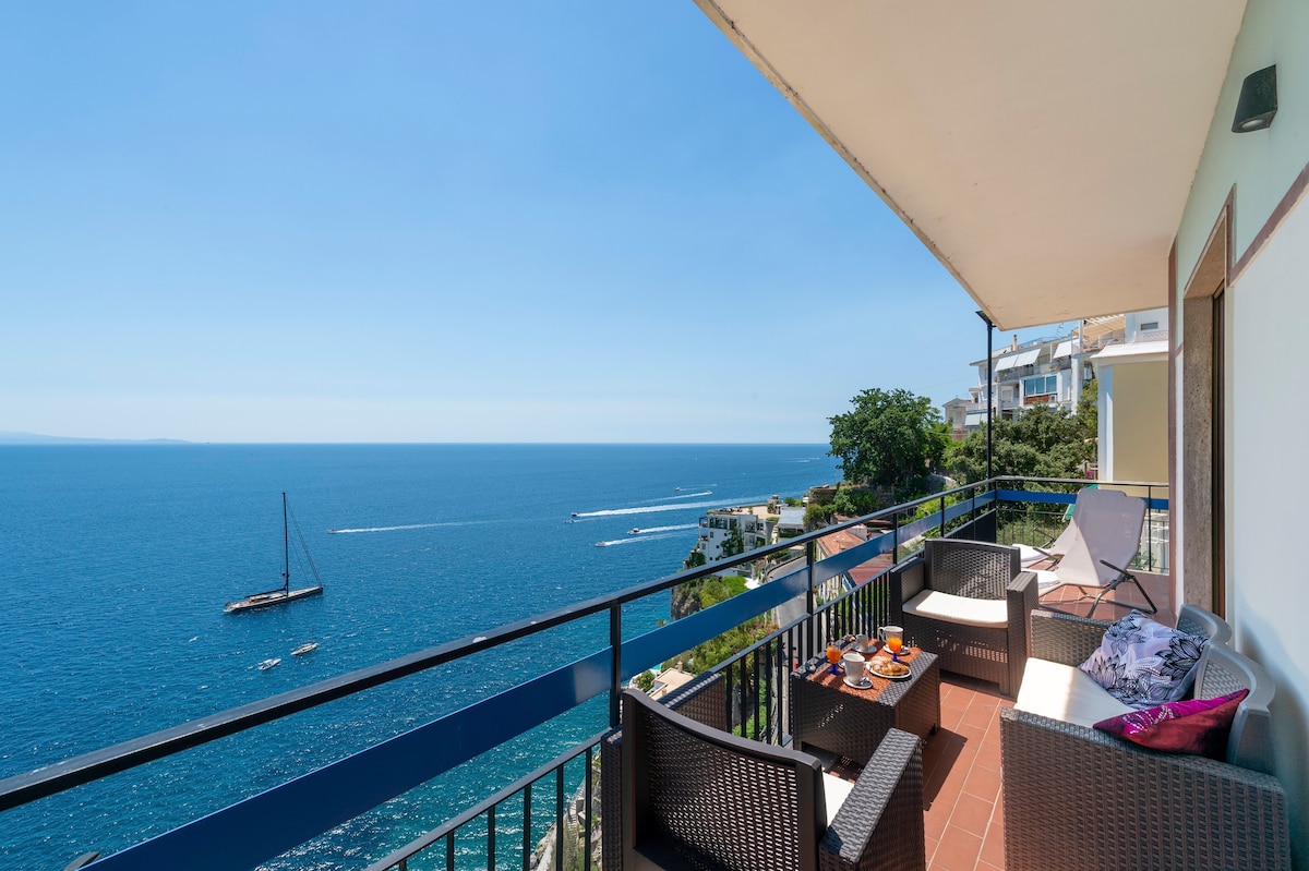 A balcony offers stunning views of the azure sea, equipped with comfortable outdoor seating. The serene setting is complemented by a table featuring refreshments, inviting relaxation while enjoying the coastal panorama.