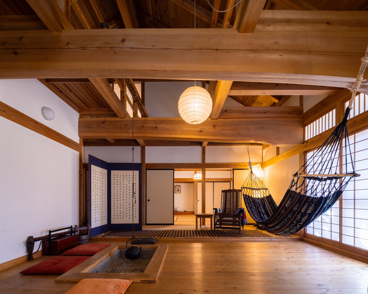 A spacious interior showcases traditional wooden beams and soft lighting, creating a calming atmosphere. A hammock is suspended in the corner, while low seating cushions are arranged around a central table. Shoji screens blend natural light with a sense of privacy.