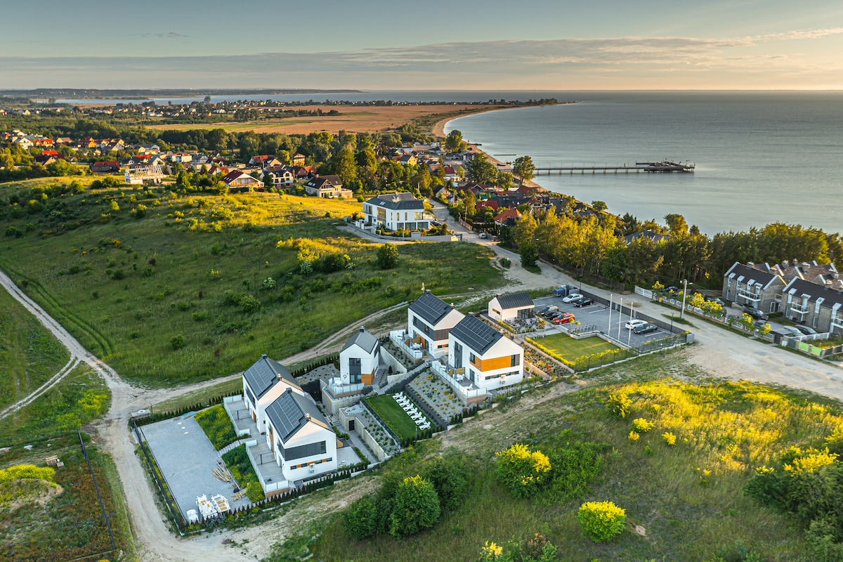 An aerial view reveals modern apartment buildings nestled among greenery, close to the coastline. The landscape features patches of grass and scattered trees, with a sandy beach visible along the horizon. A portion of the town and a pier are seen in the distance.