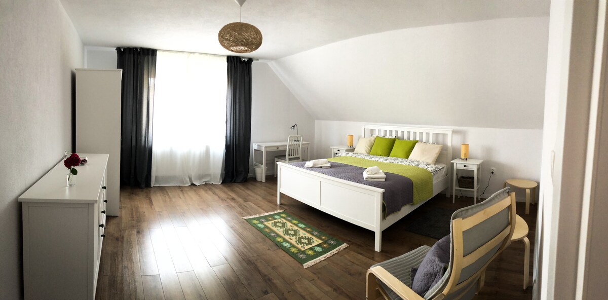 A spacious bedroom features a large bed dressed with neutral and green linens. Natural light filters through a large window, complemented by dark curtains. A table and chairs provide a workspace, while a decorative rug adds a touch of warmth to the wooden floor.