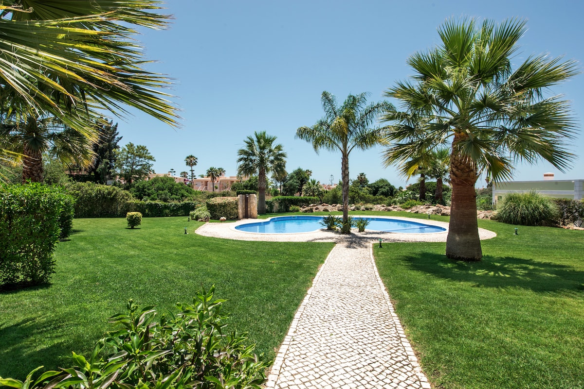 A well-maintained garden is shown, featuring a circular swimming pool surrounded by lush greenery and palm trees. A winding cobblestone pathway leads towards the pool, providing access to the tranquil outdoor space. The sky above is clear and bright, enhancing the inviting atmosphere.