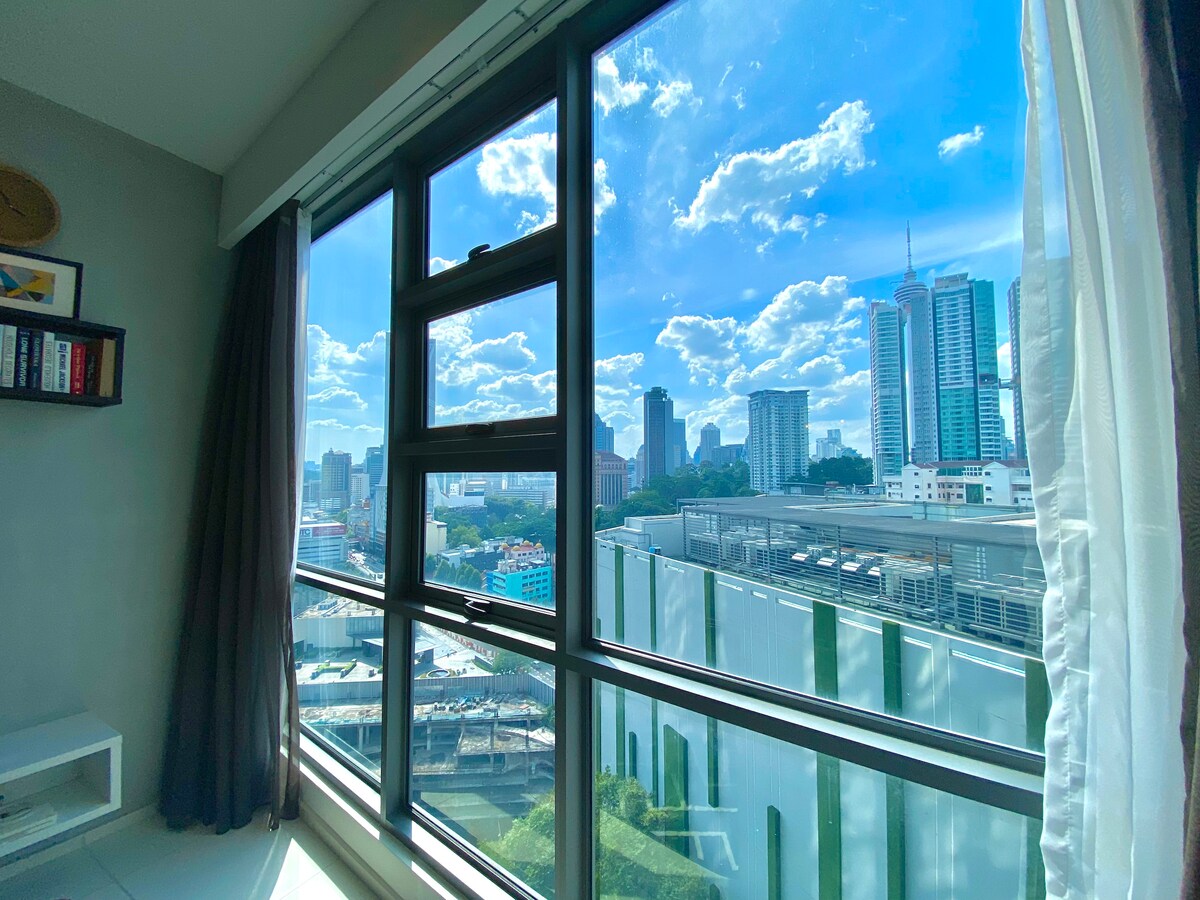Large windows provide a panoramic view of the Kuala Lumpur skyline, showcasing a blend of modern skyscrapers against a bright blue sky adorned with fluffy clouds. Natural light fills the space, creating a bright and airy environment.