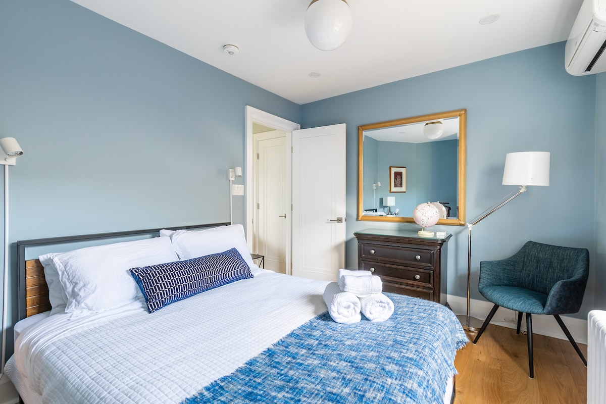 A well-lit bedroom features a spacious bed dressed in crisp white linens, complemented by a decorative blue throw. A sleek mirror and a wooden dresser are visible, along with a cozy chair and a lamp in the corner, enhancing the room's functionality.
