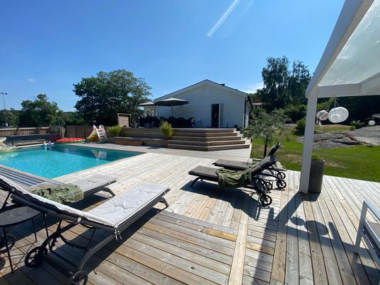 A pool area features a clear blue pool surrounded by a wooden deck. Several lounge chairs are positioned nearby. In the background, a light-colored building is visible, complemented by shaded seating and greenery, all under a clear sky.