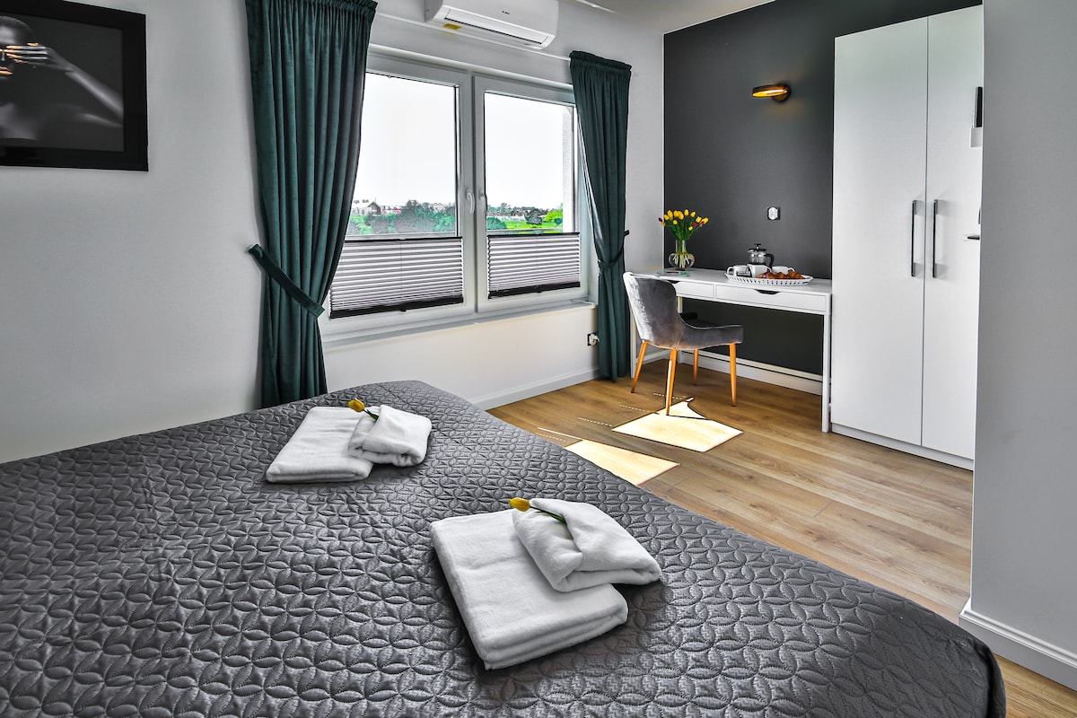 A well-appointed bedroom features a spacious bed adorned with folded towels and small decorative flowers. Two large windows allow natural light to fill the space, complemented by sheer curtains. A stylish desk and chair are positioned near a contemporary wardrobe and a wall-mounted television.