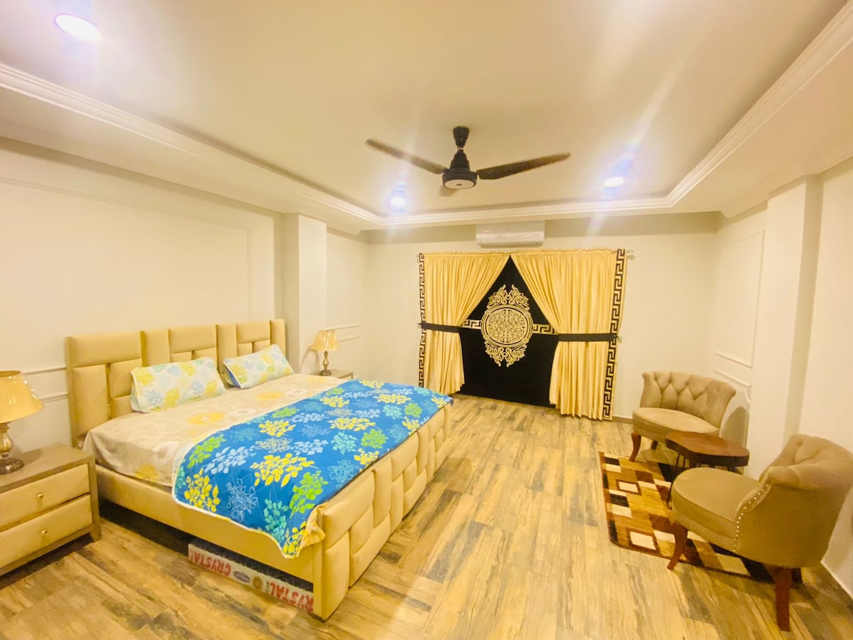 The spacious bedroom features a large bed with a patterned blue and green blanket. Two bedside lamps are positioned on either side of the bed, while a pair of accent chairs and a small table are situated nearby. Floor-to-ceiling curtains frame a window, providing a soft, warm ambiance.