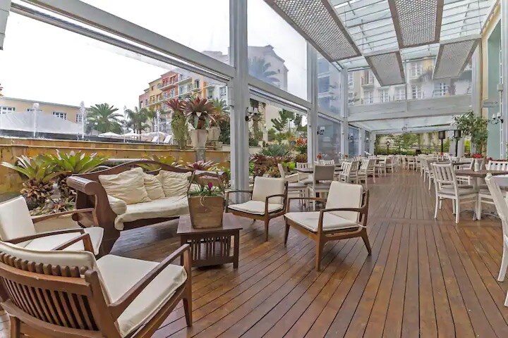 An inviting terrace is shown, featuring a wooden floor and surrounded by lush greenery. Comfortable seating arrangements include white chairs with wooden frames and a cozy sofa. Large glass panels allow natural light to fill the space, creating a bright and airy atmosphere.