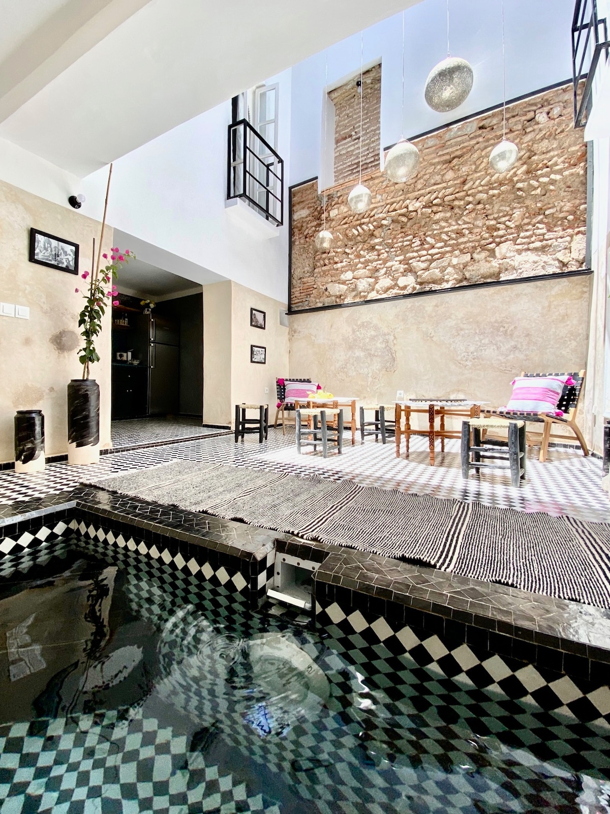 An indoor space features a reflective pool with black and white patterned tiles. Natural light floods the area, highlighting a textured stone wall. Two dining tables are arranged with chairs, and decorative cushions add color, creating a warm and inviting environment.