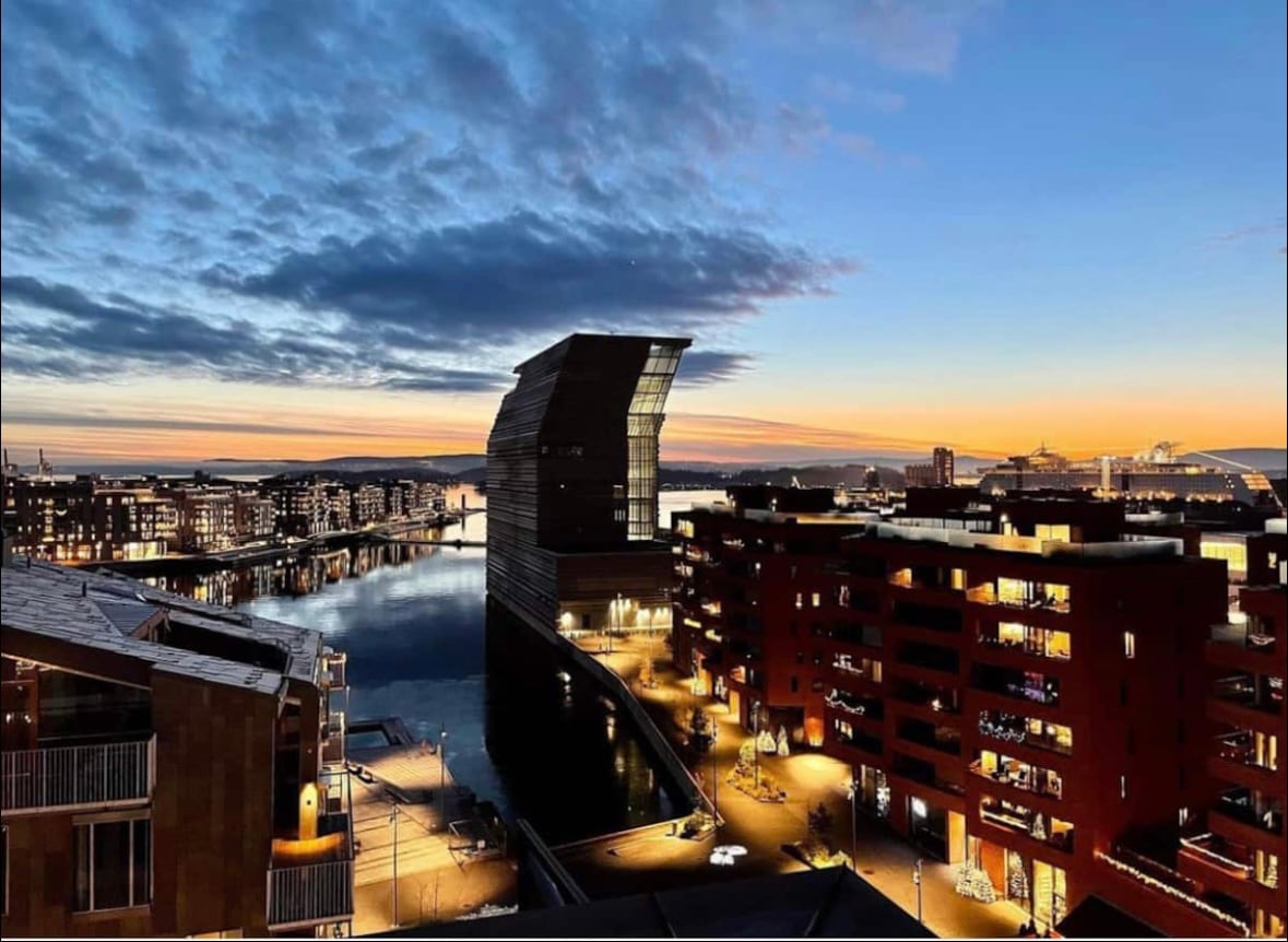 An evening view of Barcode/Bjørvika reveals a skyline filled with modern architecture. Buildings reflect warm lights along the water's edge, while soft colors of the sunset enhance the serene ambiance. The scene captures a blend of urban life and natural beauty.