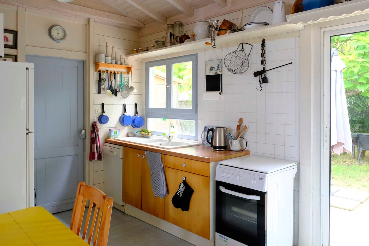 A functional kitchen is depicted with wooden cabinets, a countertop, and wall-mounted shelves holding various kitchenware. A window above the sink allows natural light, while a kettle and cooking utensils are neatly arranged. The adjoining door leads to an outdoor space.