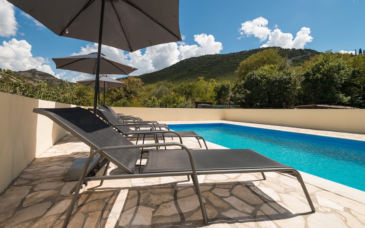 Poolside loungers are positioned under large umbrellas, overlooking a clear blue swimming pool. The surrounding area features lush greenery and distant hills beneath a bright sky filled with scattered clouds.