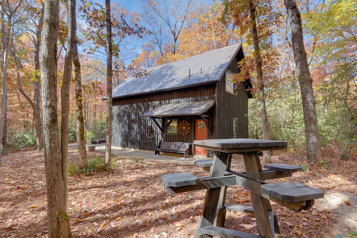 The barn loft guest house is surrounded by autumn foliage, with a combination of brown and orange leaves covering the ground. A wooden picnic table is positioned in the foreground, providing a space for outdoor meals. The building features a dark exterior and large windows.