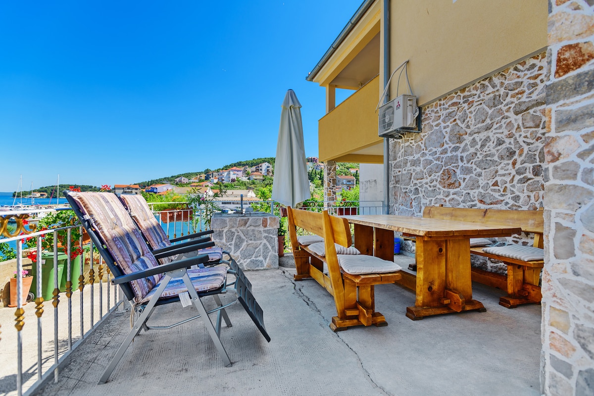 An outdoor terrace is shown with a wooden dining table and benches, complemented by two reclining lounge chairs. A large umbrella provides shade, while a stone wall adds texture. Scenic views of a marina and surrounding greenery can be seen in the background.
