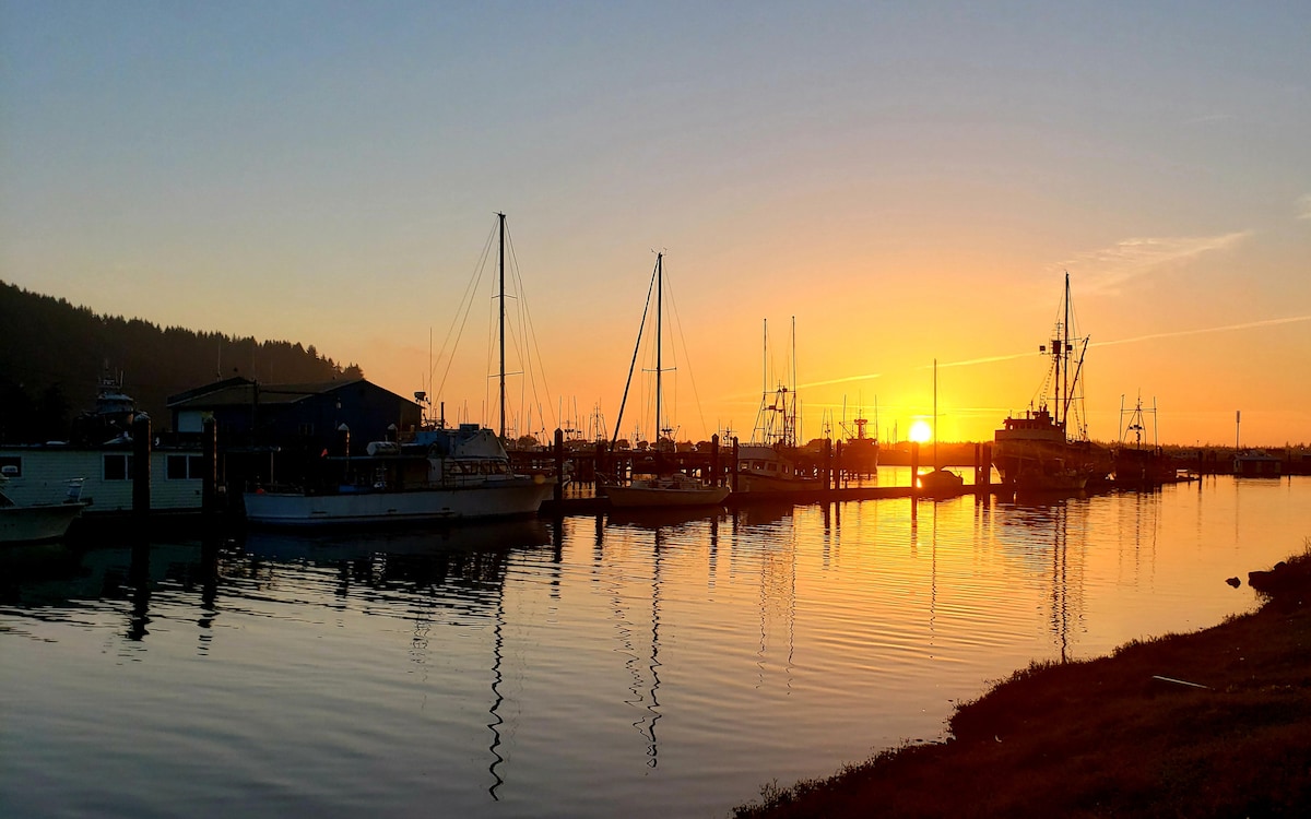 A serene harbor scene captures boats docked along calm waters, reflecting the vibrant orange and yellow hues of a sunset. Silhouettes of masts and structures outline the horizon against a backdrop of hills, creating a peaceful atmosphere by the water.